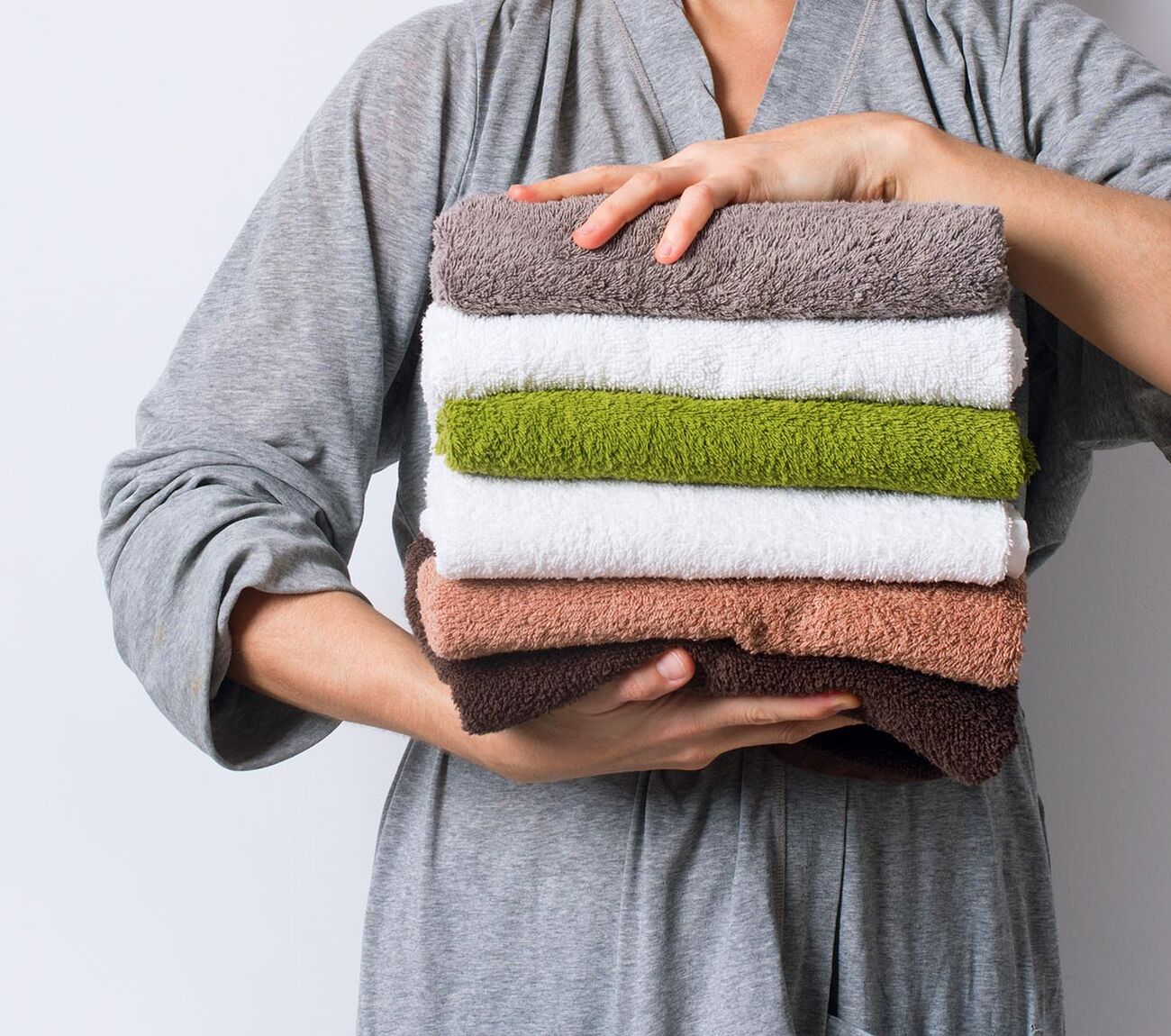 Torsobild einer Person in grauem Bademantel, die einen Stapel gefalteter Handtücher in neutralen und grünen Tönen hält.