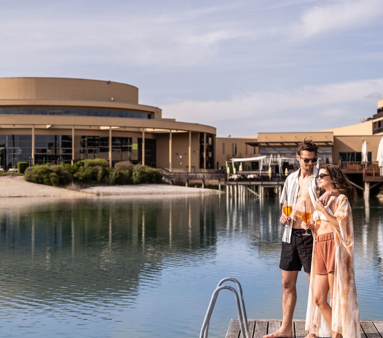 Pärchen steht umarmt auf der St. Martins Lodge See Spa Terrasse mit Cocktails in der Hand und genießt den Ausblick auf den Badesee
