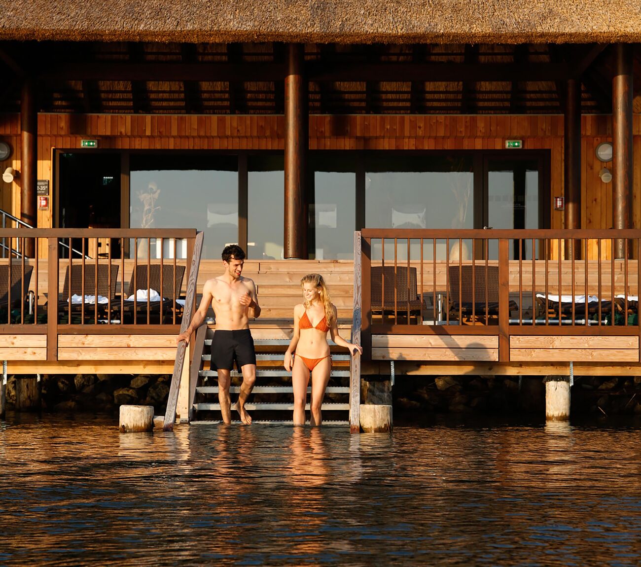 Ein Pärchen steigt von der See-Sauna über Treppen direkt in den hauseigenen Badesee der St. Martins Therme, Sonnenlicht spiegelt auf dem Wasser.