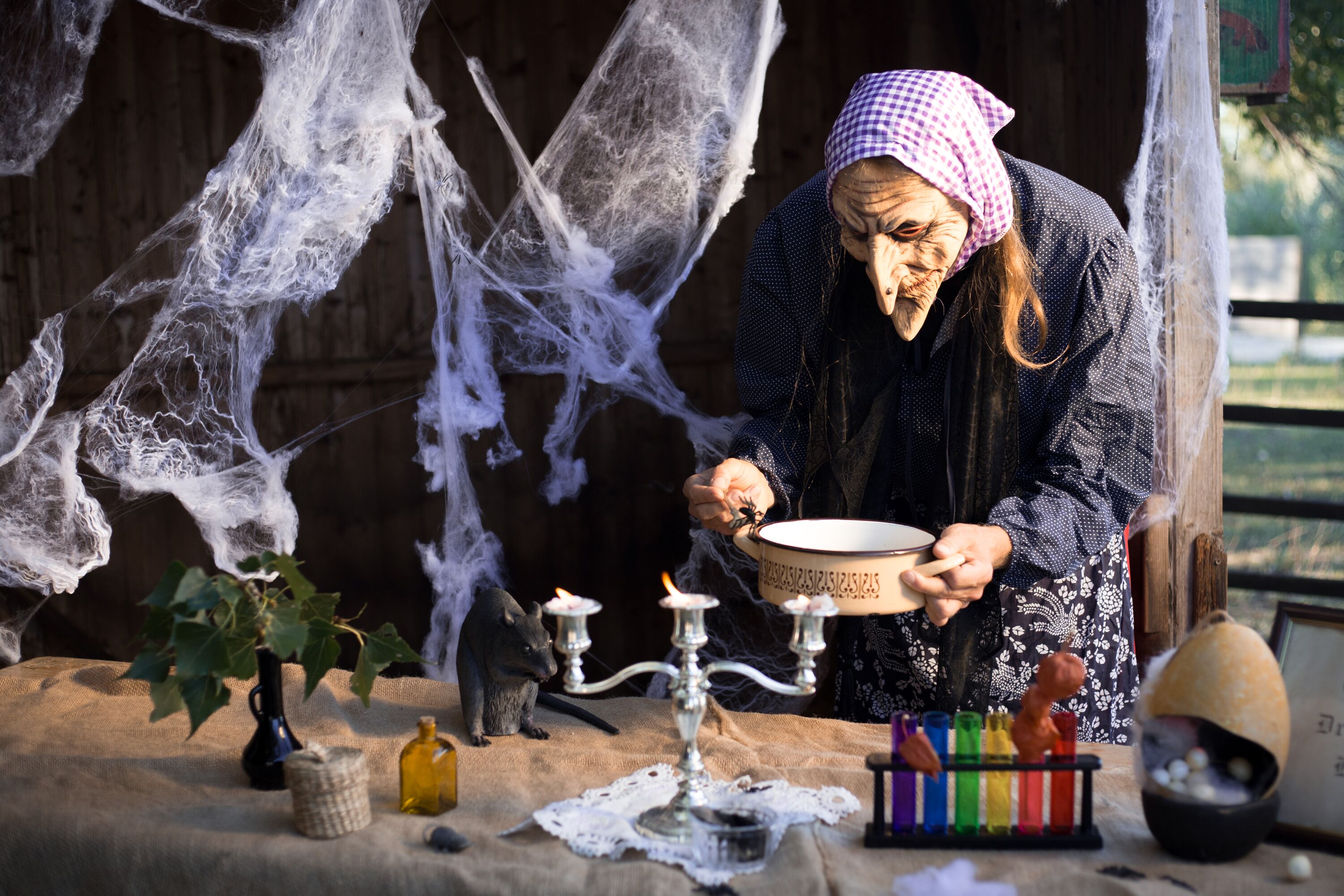 Person in Hexenmaske und Kopftuch beugt sich über Tisch mit Schale, Kerzen, Spinnennetzen, Rattenfigur und bunten Reagenzgläsern.