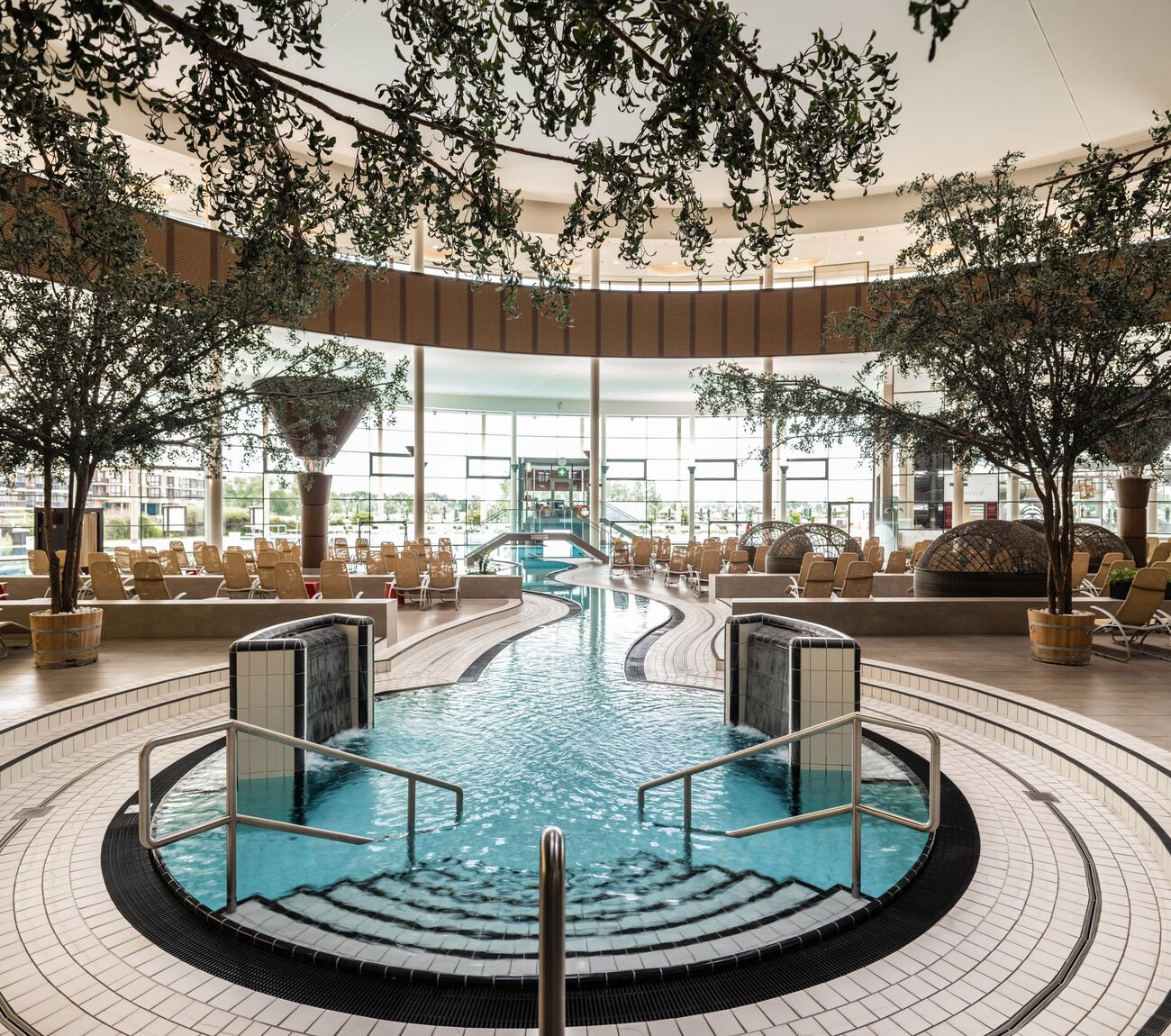 Thermenatrium mit geschwungenem Innenbecken, Treppen zum Wasser, Liegestühle und Bäumen im Hintergrund.