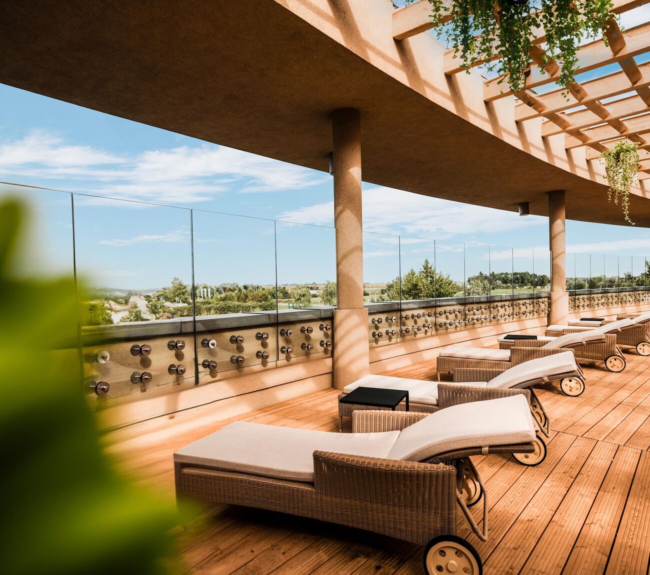 Sunset-Terrasse mit Holzdeck, Reihe von gepolsterten Liegestühlen auf Rollrädern, Glasgeländer und Pergola mit Hängepflanzen.